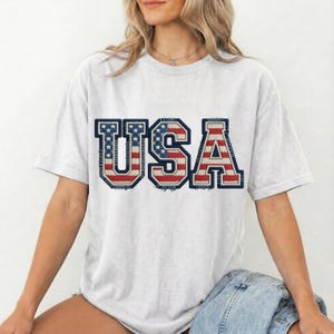 May include: A light gray t-shirt with the word "USA" in a patriotic design. The letters are in red, white, and blue, resembling the American flag. The shirt has a casual, relaxed fit.