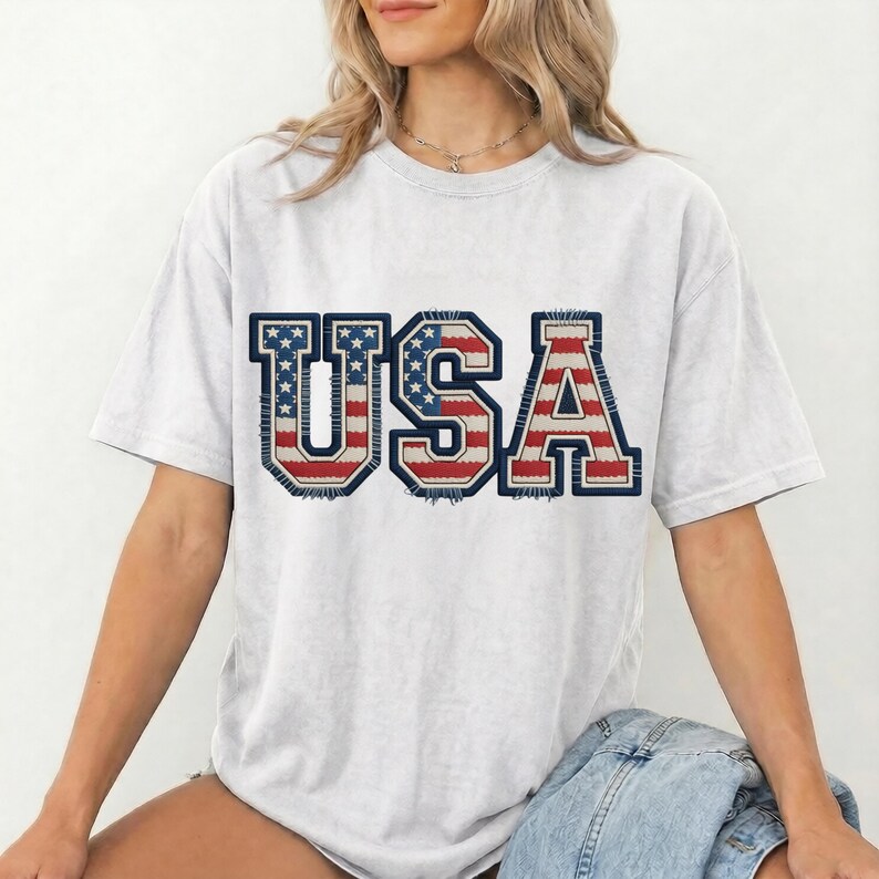 May include: A light gray t-shirt with the word "USA" in a patriotic design. The letters are in red, white, and blue, resembling the American flag. The shirt has a casual, relaxed fit.