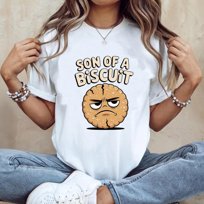 May include: White t-shirt featuring the phrase "SON OF A BISCUIT" above a cartoon biscuit with an angry face. The biscuit is round, with a textured surface and a grumpy expression. The shirt is a casual, everyday wear item.