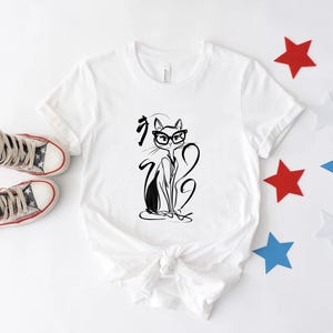 May include: White t-shirt featuring a black and white illustration of a cat wearing glasses. The cat design is accompanied by Japanese characters. The shirt is paired with high-top sneakers and star-shaped decorations in red, white, and blue.