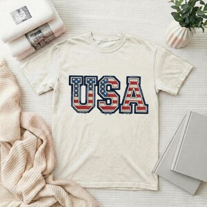 May include: Cream-colored t-shirt with the word "USA" in a patriotic design. The letters are filled with the American flag's stars and stripes, outlined in navy blue. The shirt is displayed on a white surface.