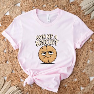 May include: A light pink t-shirt with the phrase "SON OF A BISCUIT" in a playful font. Below the text is a cartoon biscuit with an angry expression. The shirt is folded and placed on a woven rug.
