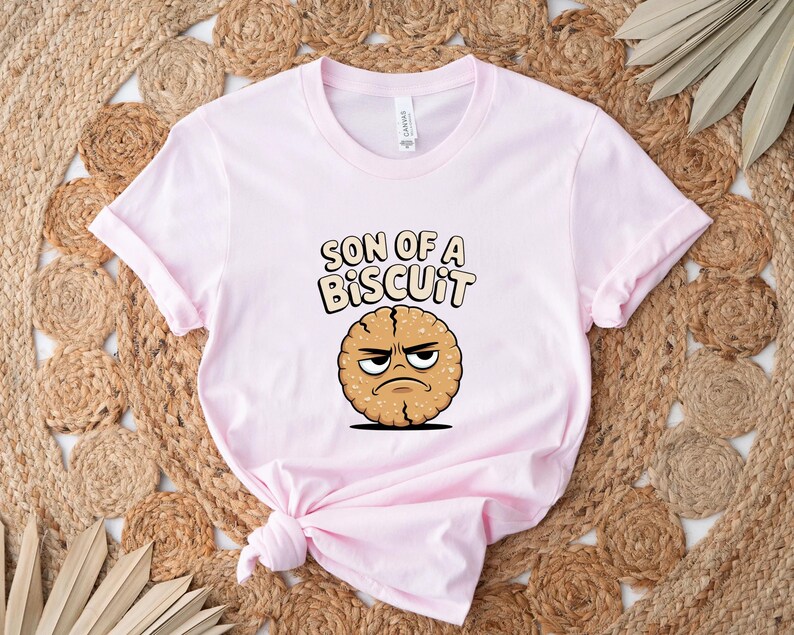 May include: A light pink t-shirt with the phrase "SON OF A BISCUIT" in a playful font. Below the text is a cartoon biscuit with an angry expression. The shirt is folded and placed on a woven rug.