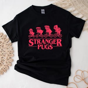 May include: Black t-shirt featuring a pink graphic of four pugs riding bicycles, above the text "STRANGER PUGS" in pink. The design is reminiscent of a popular science fiction series. A casual, fun tee.