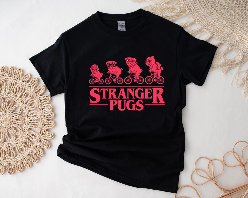 May include: Black t-shirt featuring a pink graphic of four pugs riding bicycles, above the text "STRANGER PUGS" in pink. The design is reminiscent of a popular science fiction series. A casual, fun tee.