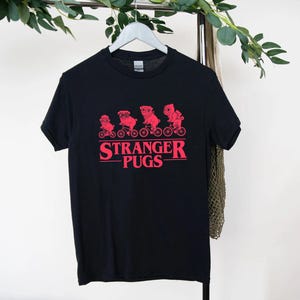May include: Black t-shirt with a red graphic of pugs riding bicycles and the text "Stranger Pugs". The design features four pugs on bikes, with the text below. The shirt is hanging on a white hanger.