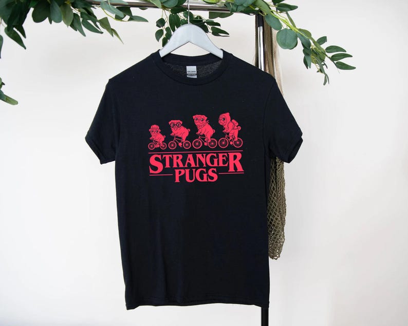 May include: Black t-shirt with a red graphic of pugs riding bicycles and the text "Stranger Pugs". The design features four pugs on bikes, with the text below. The shirt is hanging on a white hanger.