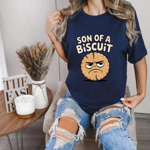 May include: Navy blue t-shirt with the text "SON OF A BISCUIT" above a cartoon biscuit with an angry face. The shirt is a crew neck and is being worn.