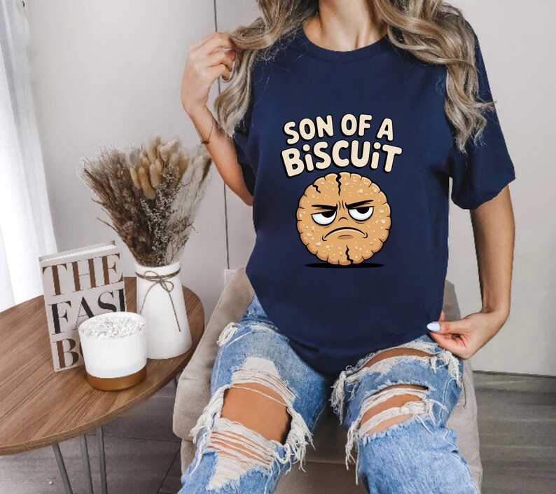May include: Navy blue t-shirt with the text "SON OF A BISCUIT" above a cartoon biscuit with an angry face. The shirt is a crew neck and is being worn.