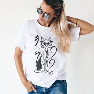 May include: White t-shirt featuring a black and white illustration of a cat wearing glasses. The cat illustration has a minimalist design with Japanese characters. The t-shirt is made of a soft material.
