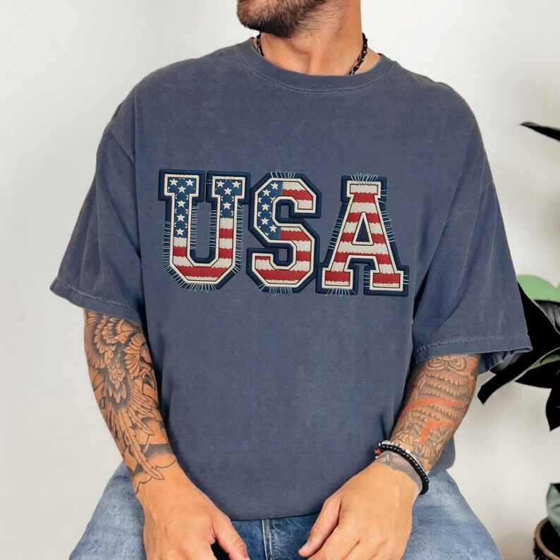 May include: A faded blue t-shirt with the word "USA" prominently displayed across the chest. The letters are designed with a patriotic theme, featuring the American flag's red and white stripes and blue stars.