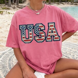 May include: A pink t-shirt with the word "USA" in a bold, patriotic design. The letters are outlined in navy blue with a red, white, and blue flag pattern. The shirt is worn with white sneakers.