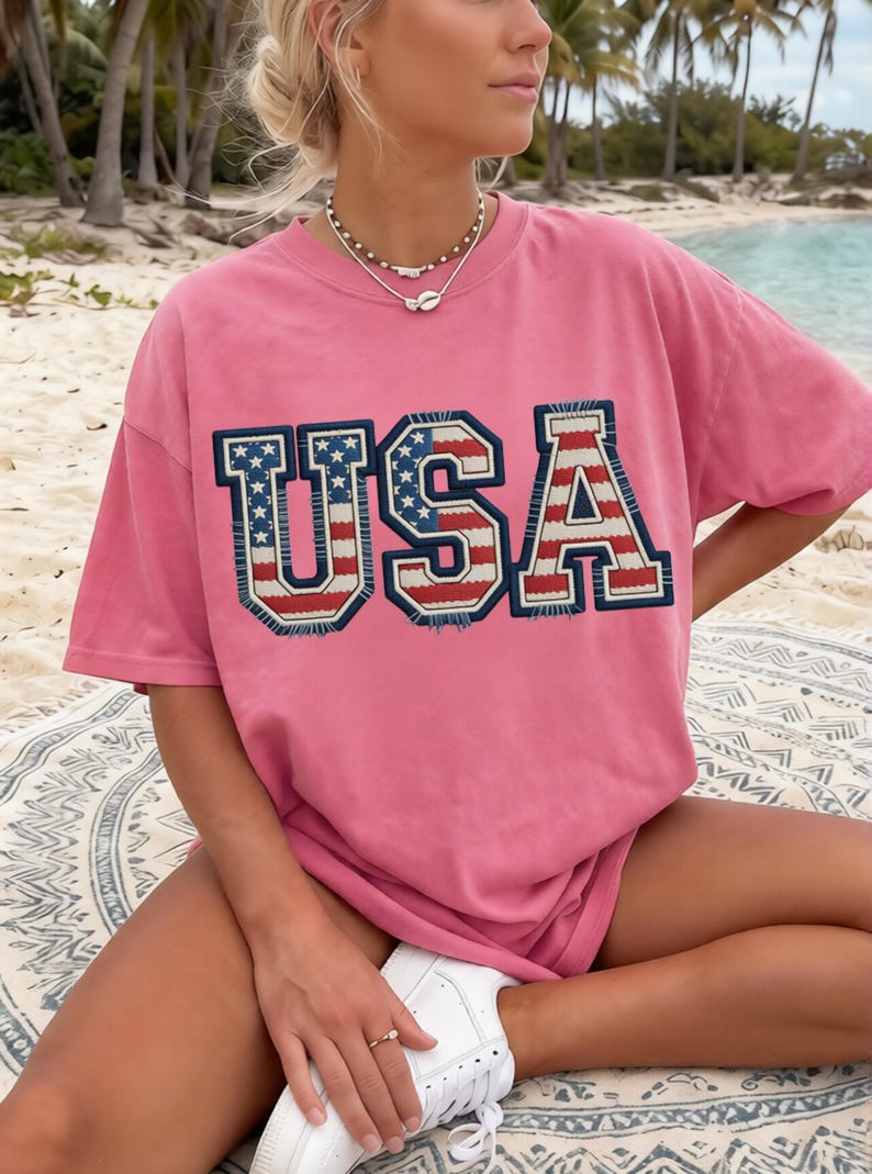 May include: A pink t-shirt with the word "USA" in a bold, patriotic design. The letters are outlined in navy blue with a red, white, and blue flag pattern. The shirt is worn with white sneakers.