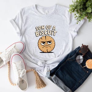 May include: A light gray t-shirt with the text "SON OF A BISCUIT" above a cartoon biscuit with an angry face. White sneakers with red trim, sunglasses, and a lollipop are also visible.