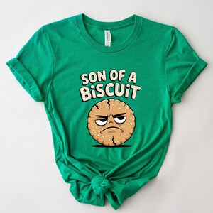 May include: A bright green t-shirt featuring the phrase "SON OF A BISCUIT" above a cartoon biscuit with an angry expression. The biscuit is light brown with a cracked texture and a black outline.