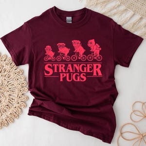May include: Burgundy t-shirt with a pink graphic of pugs riding bicycles above the text "STRANGER PUGS". The design features four pugs, each on a bicycle, with the text in a bold, stylized font.