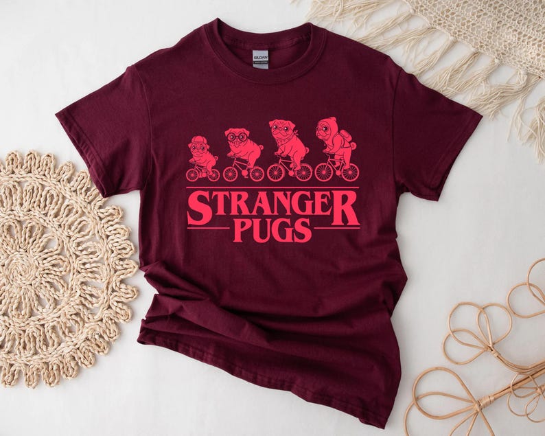 May include: Burgundy t-shirt with a pink graphic of pugs riding bicycles above the text "STRANGER PUGS". The design features four pugs, each on a bicycle, with the text in a bold, stylized font.