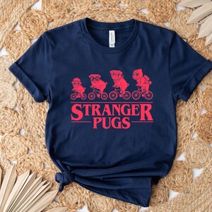 May include: Navy blue t-shirt featuring a red graphic of pugs riding bicycles, with the text "Stranger Pugs" in a red, stylized font. The shirt is made of a soft material and has a classic crew neck.