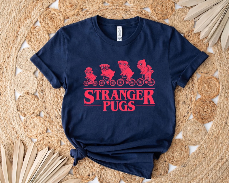 May include: Navy blue t-shirt featuring a red graphic of pugs riding bicycles, with the text "Stranger Pugs" in a red, stylized font. The shirt is made of a soft material and has a classic crew neck.
