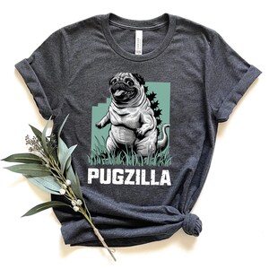 May include: A dark gray t-shirt with a graphic of a pugzilla, a pug with a Godzilla-like appearance, standing on a green patch of grass. The text "PUGZILLA" is printed in white below the graphic.