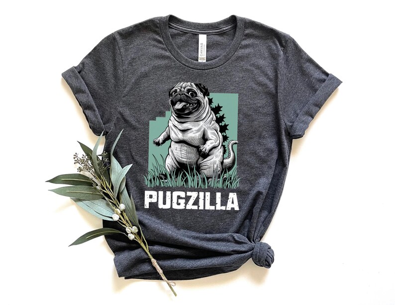 May include: A dark gray t-shirt with a graphic of a pugzilla, a pug with a Godzilla-like appearance, standing on a green patch of grass. The text "PUGZILLA" is printed in white below the graphic.