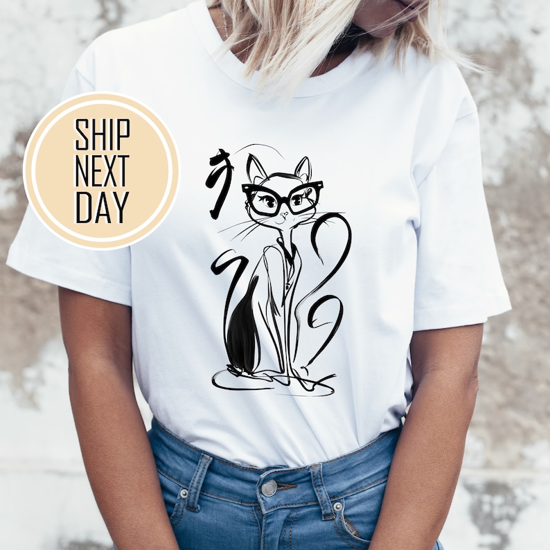 May include: White t-shirt featuring a black and white illustration of a cat wearing glasses. The cat is drawn in a minimalist style with long, flowing lines. The shirt has a classic crew neck and short sleeves.