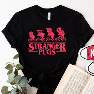 May include: Black t-shirt featuring a red graphic of four pugs riding bicycles, with the text "Stranger Pugs" in a retro font. The design is centered on the shirt. The shirt is paired with red sneakers and a book.