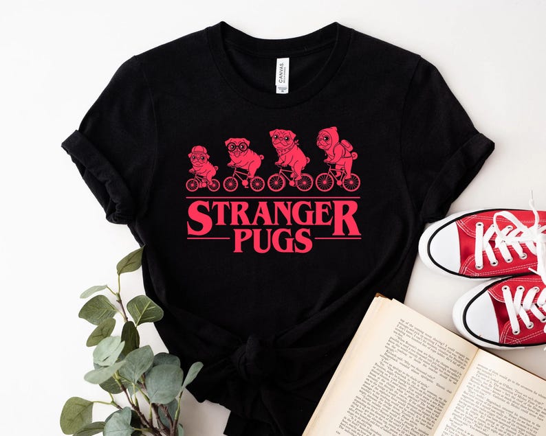 May include: Black t-shirt featuring a red graphic of four pugs riding bicycles, with the text "Stranger Pugs" in a retro font. The design is centered on the shirt. The shirt is paired with red sneakers and a book.
