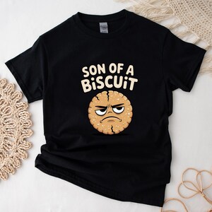 May include: Black t-shirt with the text "SON OF A BISCUIT" above a cartoon biscuit with an angry face. The biscuit is light brown with a darker brown outline and details.