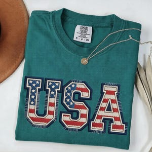 May include: Teal-colored t-shirt with the word "USA" in a patriotic design. The letters are outlined in navy blue and filled with the American flag pattern. A gold pendant necklace and a brown hat are also visible.