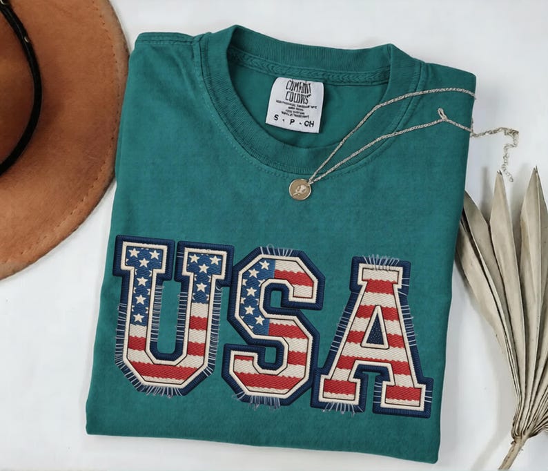 May include: Teal-colored t-shirt with the word "USA" in a patriotic design. The letters are outlined in navy blue and filled with the American flag pattern. A gold pendant necklace and a brown hat are also visible.