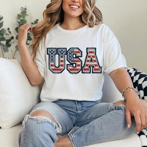 May include: White t-shirt with the word "USA" in a patriotic design. The letters are outlined in navy blue, with a red and white striped pattern and blue stars filling the letters. The shirt is worn with blue jeans.