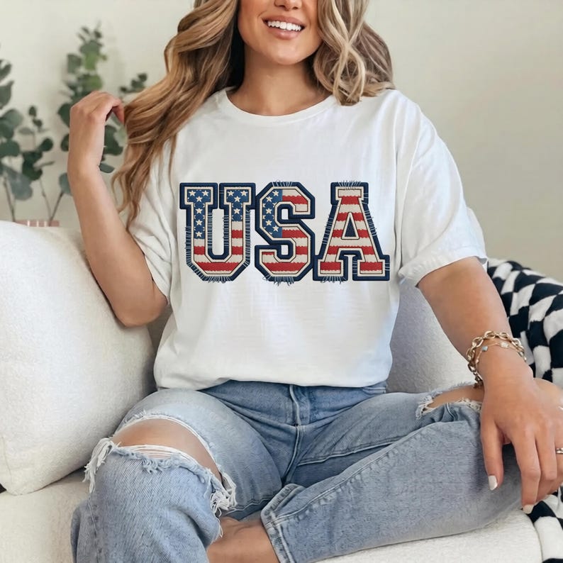 May include: White t-shirt with the word "USA" in a patriotic design. The letters are outlined in navy blue, with a red and white striped pattern and blue stars filling the letters. The shirt is worn with blue jeans.