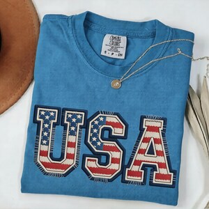May include: A blue t-shirt with the word "USA" embroidered across the front in a patriotic design. The letters feature the American flag's stars and stripes. A gold necklace with a pendant rests on the shirt.