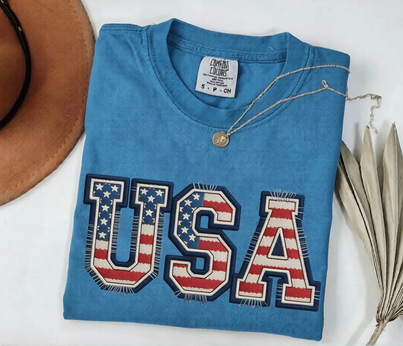 May include: A blue t-shirt with the word "USA" embroidered across the front in a patriotic design. The letters feature the American flag's stars and stripes. A gold necklace with a pendant rests on the shirt.