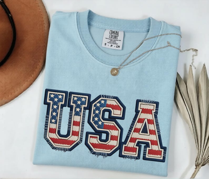 May include: A light blue t-shirt with the word "USA" embroidered on the front in red, white, and blue. The shirt has a crew neck and short sleeves. A silver necklace with a small pendant is draped across the shirt.
