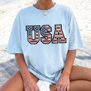 May include: Light blue t-shirt with the word "USA" in a patriotic design. The letters are in red, white, and blue, with a distressed, vintage look. The shirt is short-sleeved and appears to be made of a soft material.