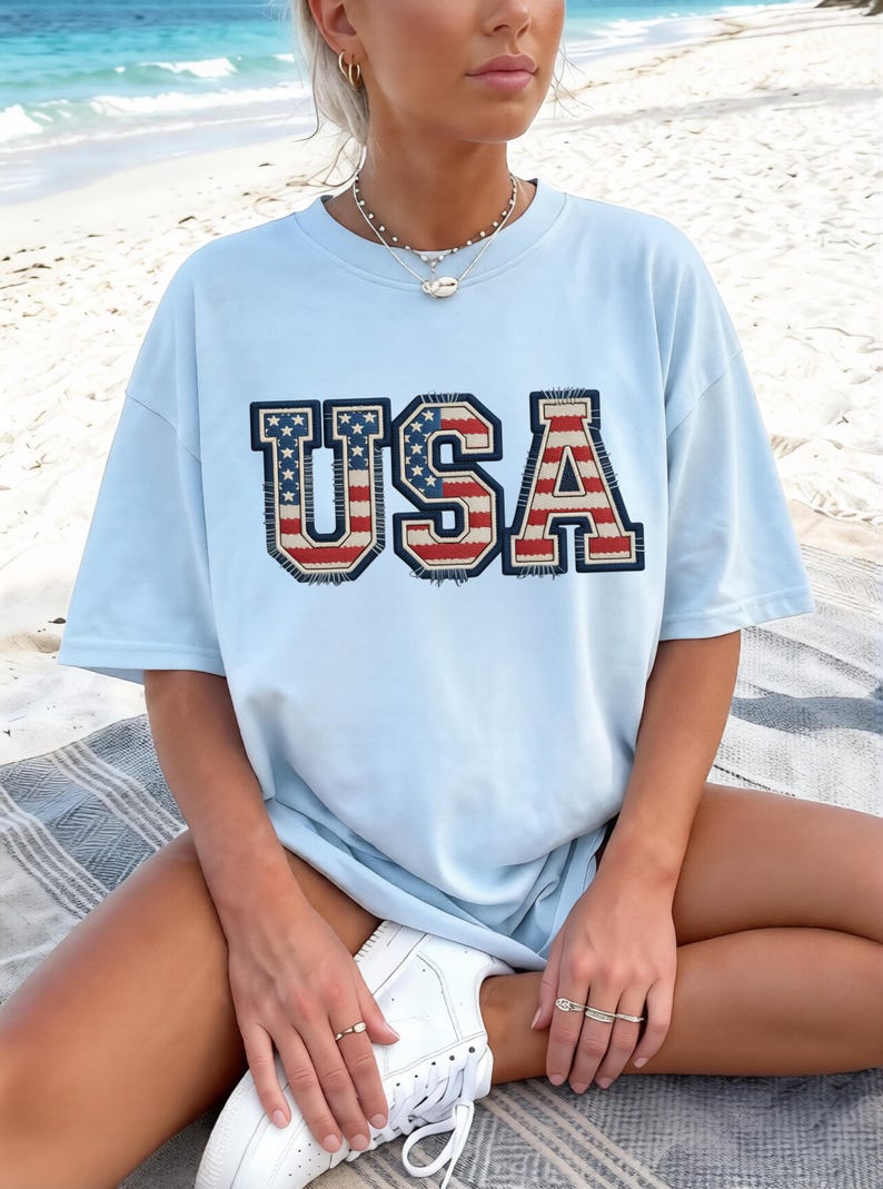 May include: Light blue t-shirt with the word "USA" in a patriotic design. The letters are in red, white, and blue, with a distressed, vintage look. The shirt is short-sleeved and appears to be made of a soft material.