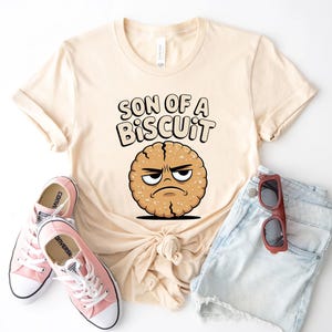 May include: Cream-colored t-shirt with the phrase "SON OF A BISCUIT" and a cartoon biscuit character with an angry expression. The shirt is paired with light blue denim shorts, pink sneakers, and red sunglasses.