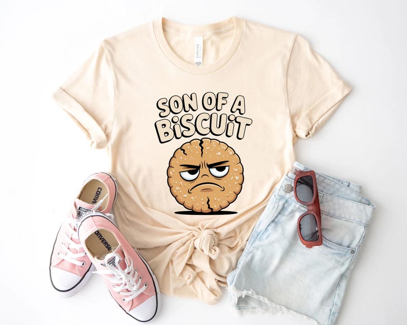 May include: Cream-colored t-shirt with the phrase "SON OF A BISCUIT" and a cartoon biscuit character with an angry expression. The shirt is paired with light blue denim shorts, pink sneakers, and red sunglasses.