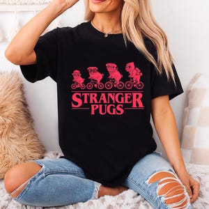May include: Black t-shirt featuring a pink graphic of pugs riding bicycles, with the text "STRANGER PUGS" in a retro font. The design is reminiscent of a popular science fiction series. The shirt is made of a soft material.