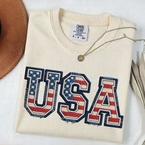 May include: Cream-colored sweatshirt with the word "USA" in a patriotic design. The letters are outlined in navy blue with red and white stripes and blue stars. A silver necklace and a brown hat are also visible.