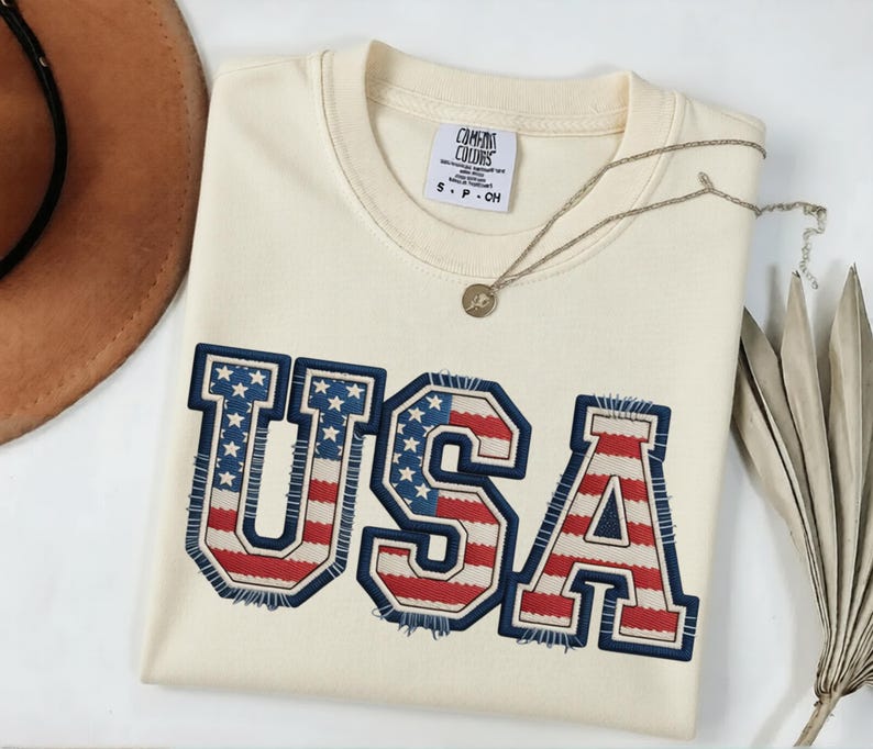 May include: Cream-colored sweatshirt with the word "USA" in a patriotic design. The letters are outlined in navy blue with red and white stripes and blue stars. A silver necklace and a brown hat are also visible.