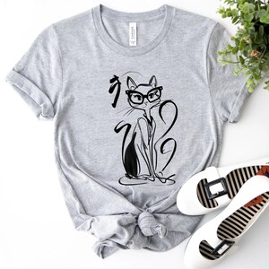 May include: A light gray t-shirt featuring a black and white illustration of a cat wearing glasses. The cat design is accompanied by Japanese characters. White flats with black and white striped soles are also visible.