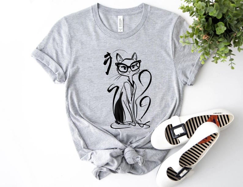 May include: A light gray t-shirt featuring a black and white illustration of a cat wearing glasses. The cat design is accompanied by Japanese characters. White flats with black and white striped soles are also visible.