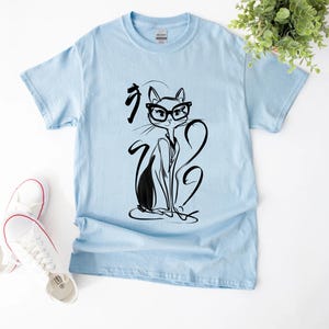 May include: Light blue t-shirt featuring a black and white illustration of a cat wearing glasses. The cat is drawn in a minimalist style with long, flowing lines. The shirt is paired with white sneakers.