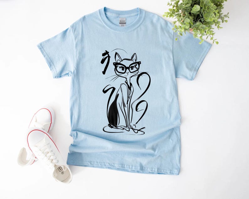 May include: Light blue t-shirt featuring a black and white illustration of a cat wearing glasses. The cat is drawn in a minimalist style with long, flowing lines. The shirt is paired with white sneakers.