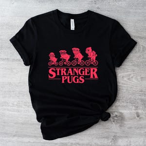 May include: Black t-shirt featuring a red graphic of pugs riding bicycles, with the text "Stranger Pugs" in a matching red font. The design is centered on the front of the shirt. The shirt is made of a soft material.