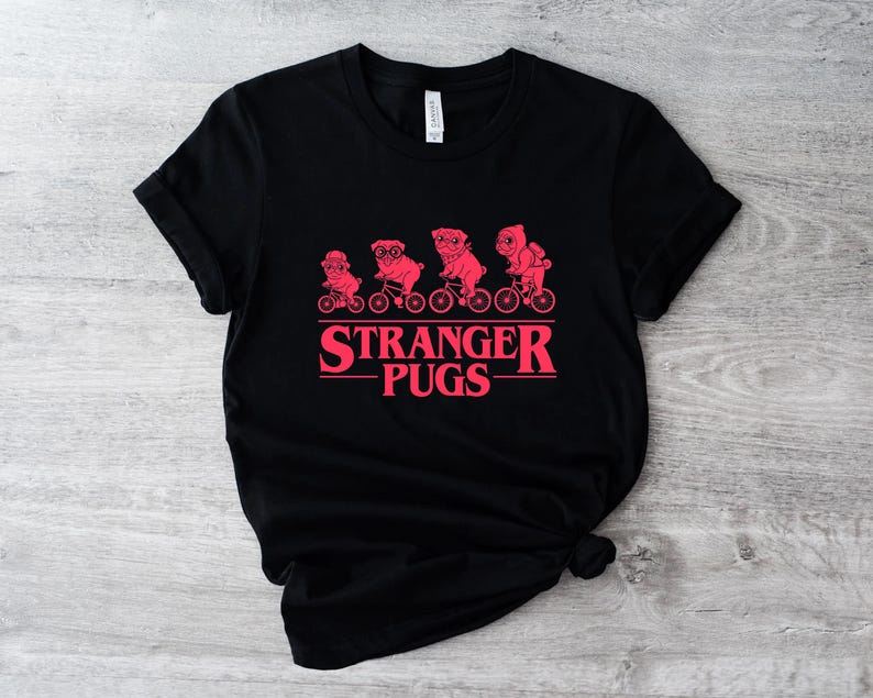 May include: Black t-shirt featuring a red graphic of pugs riding bicycles, with the text "Stranger Pugs" in a matching red font. The design is centered on the front of the shirt. The shirt is made of a soft material.