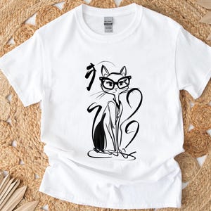 May include: White t-shirt featuring a black and white illustration of a cat wearing glasses. The cat is drawn in a minimalist style with long, flowing lines. The shirt also has Japanese characters above the cat.